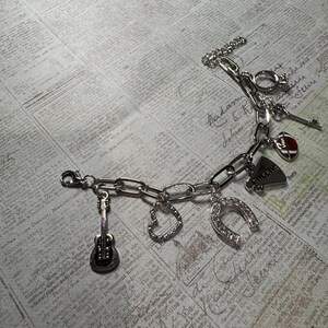 Puede incluir: Pulsera de abalorios plateada con dijes de guitarra, coraz&oacute;n, herradura, meg&aacute;fono y f&uacute;tbol. La pulsera tiene un cierre de palanca y se muestra sobre un fondo de peri&oacute;dico vintage.