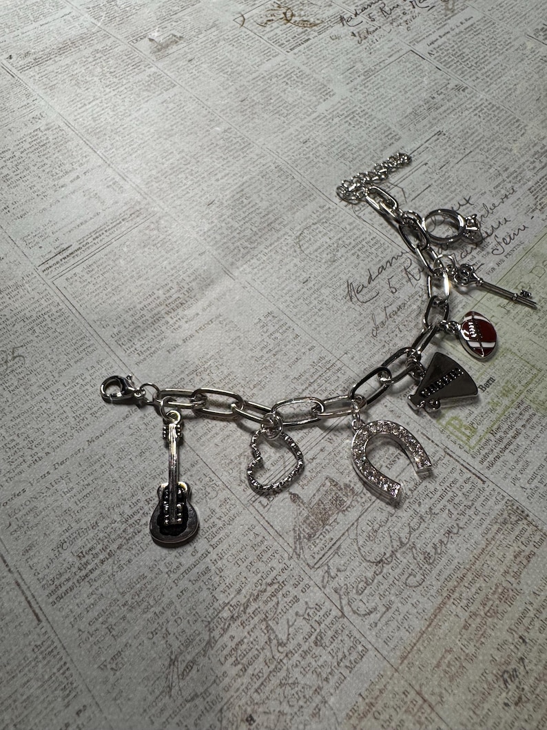 Puede incluir: Pulsera de abalorios plateada con dijes de guitarra, coraz&oacute;n, herradura, meg&aacute;fono y f&uacute;tbol. La pulsera tiene un cierre de palanca y se muestra sobre un fondo de peri&oacute;dico vintage.