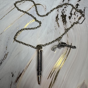May include: A necklace with a silver bullet pendant and a small gun charm. The chain is a dark bronze color. The bullet pendant is silver with a gold-colored tip. The gun charm is a detailed, antique-style design.