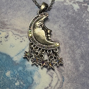 May include: Silver-toned necklace featuring a crescent moon pendant with a face design and dangling stars. The moon is embellished with iridescent rhinestones. The chain is also silver-toned.