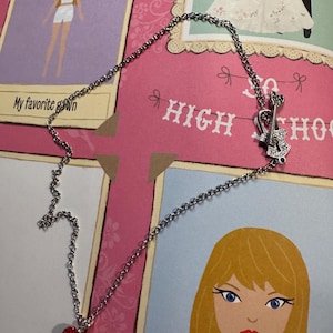 May include: A silver necklace featuring a red heart pendant and a guitar-shaped charm. The necklace is set against a pink backdrop with the words "HIGH SCHOOL" and illustrations of a bride and a cartoon girl.