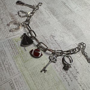 Puede incluir: Pulsera de abalorios plateada con varios dijes, incluyendo una herradura, un bal&oacute;n de f&uacute;tbol americano, una llave y un anillo. La pulsera est&aacute; hecha de cadena de eslabones y se muestra sobre un fondo de peri&oacute;dico vintage.