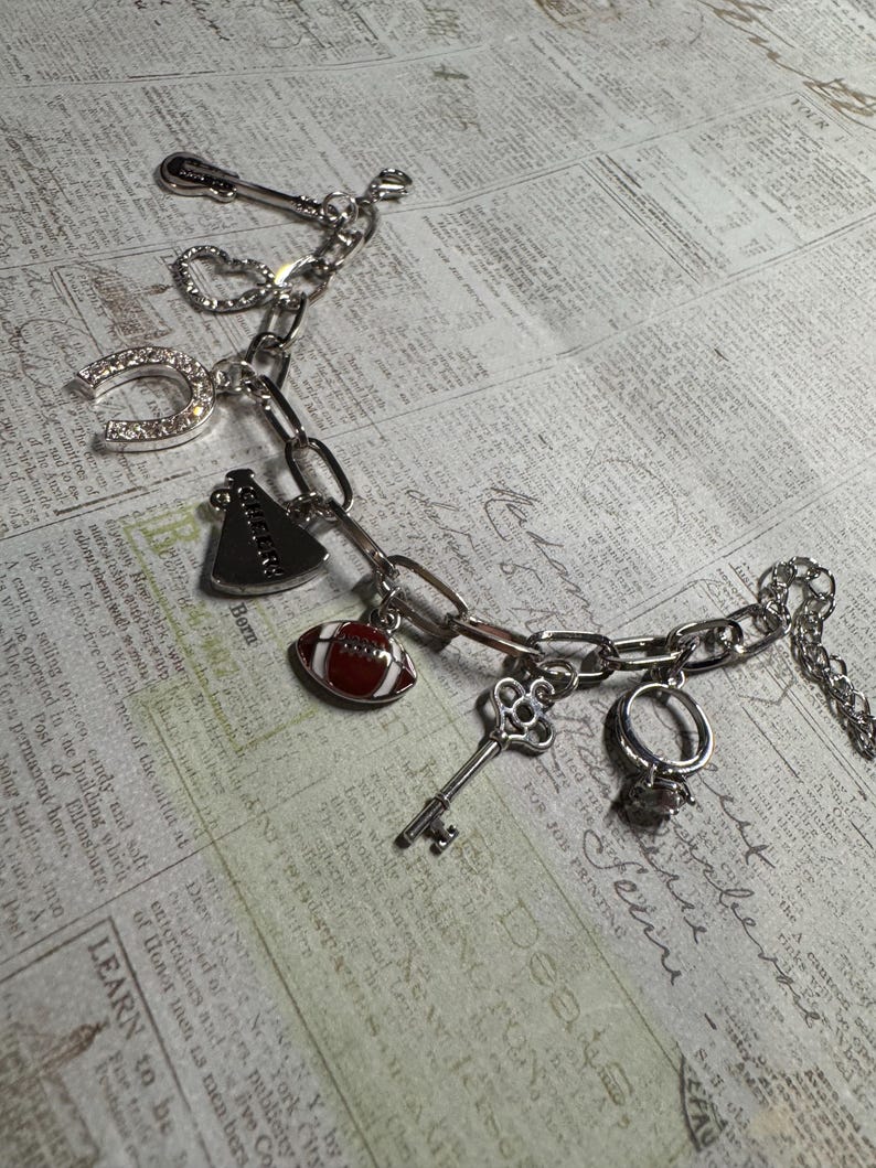 Puede incluir: Pulsera de abalorios plateada con varios dijes, incluyendo una herradura, un bal&oacute;n de f&uacute;tbol americano, una llave y un anillo. La pulsera est&aacute; hecha de cadena de eslabones y se muestra sobre un fondo de peri&oacute;dico vintage.