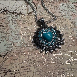 May include: Silver-toned necklace with a pendant featuring a turquoise heart-shaped stone at the center, surrounded by a dark silver floral design. The necklace rests on a vintage map background.