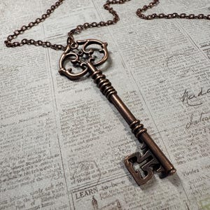 May include: A bronze-colored key pendant with an ornate design hangs from a matching chain. The key is detailed with decorative swirls and a textured handle. The necklace rests on a vintage newspaper background.