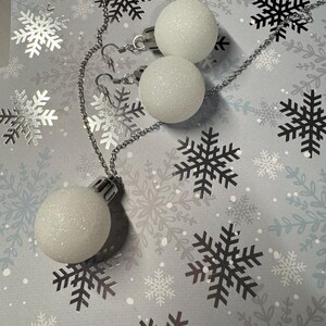 May include: A white glittery Christmas ornament necklace and matching earrings set. The necklace features a single ornament pendant, and the earrings have two ornaments. The jewelry is on a silver chain, set against a snowflake patterned background.