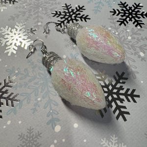 May include: A pair of Christmas light bulb earrings. The earrings are white with iridescent glitter and silver-colored metal findings. The earrings are on a gray background with snowflake patterns.