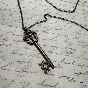 May include: A vintage-style key pendant necklace. The key is bronze-colored with ornate detailing on the head and a simple cross-shaped design on the bottom. The chain is a dark bronze color. The background is a white surface with cursive writing.