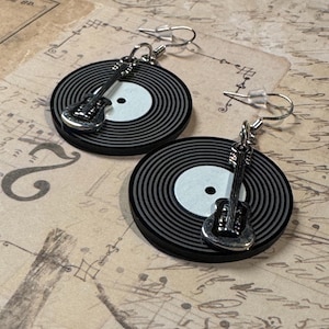 May include: A pair of earrings featuring black and gray vinyl record designs with a silver-colored guitar charm. The earrings have silver-colored ear wires. The background is a vintage-style paper.