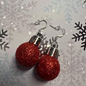 May include: A pair of festive red glitter ball earrings with silver-colored hooks. The earrings are designed to resemble miniature Christmas ornaments. The background features a light gray surface with snowflake patterns.