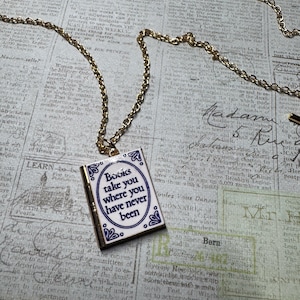 May include: A gold-toned metal chain necklace featuring a rectangular pendant shaped like a book. The pendant is white with blue decorative borders and the text "Books take you where you have never been" printed in a dark blue serif font on the front.