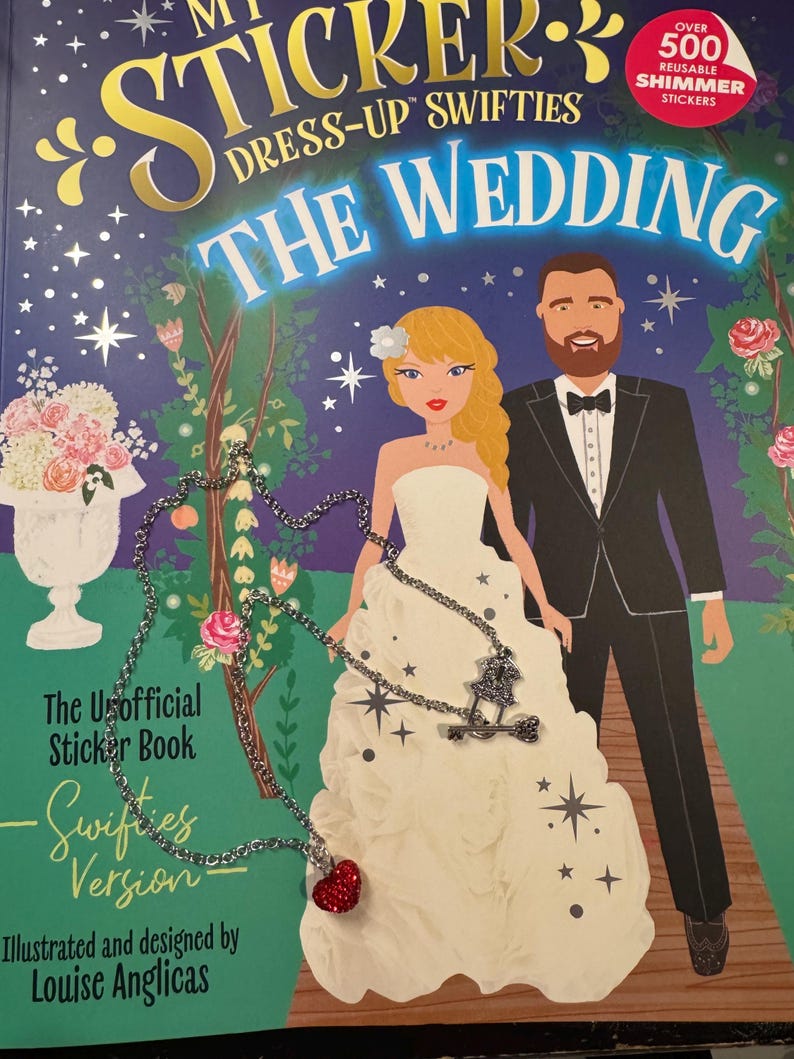 May include: A sticker book cover, "My Sticker Dress-Up Swifties: The Wedding", shows a bride and groom illustration. The cover text includes "Over 500 Reusable Shimmer Stickers." A silver necklace with a red heart pendant is in the foreground.