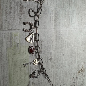 Puede incluir: Una pulsera de plata con dijes que incluye una guitarra, un coraz&oacute;n, una herradura, un velo de novia, un bal&oacute;n de f&uacute;tbol americano, una llave y un anillo. La pulsera se muestra sobre un fondo de peri&oacute;dico antiguo.