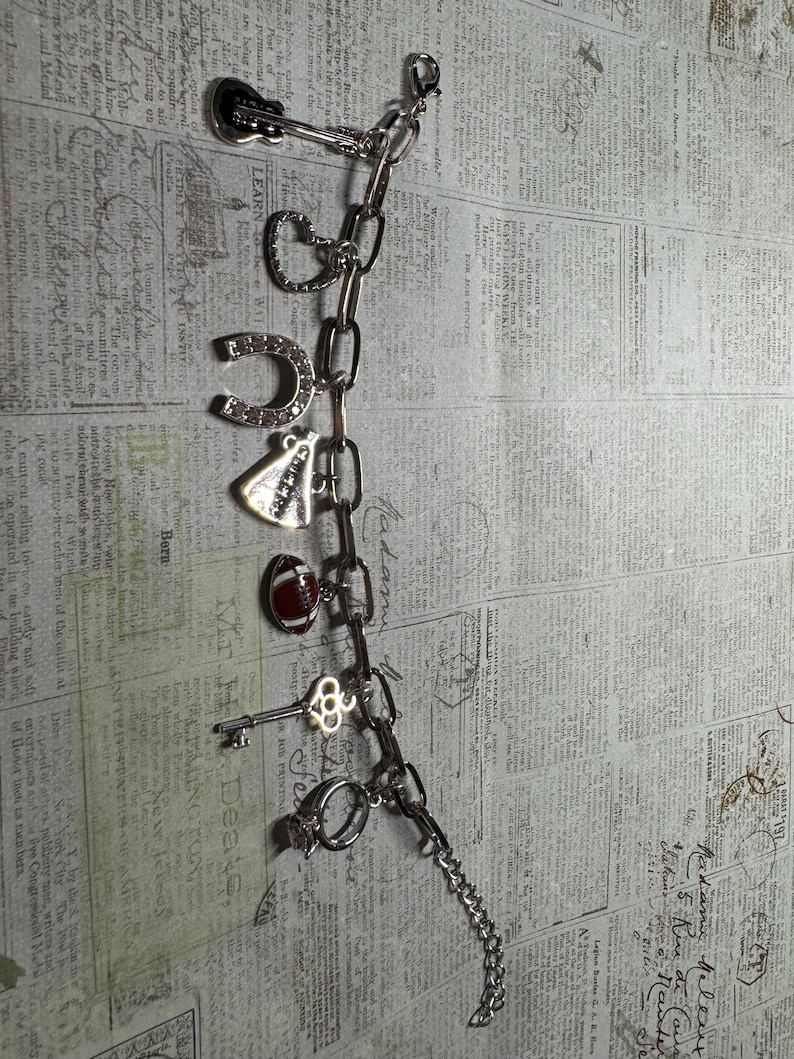 Puede incluir: Una pulsera de plata con dijes que incluye una guitarra, un coraz&oacute;n, una herradura, un velo de novia, un bal&oacute;n de f&uacute;tbol americano, una llave y un anillo. La pulsera se muestra sobre un fondo de peri&oacute;dico antiguo.