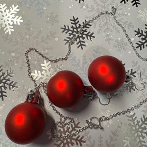 May include: A silver chain necklace with three red Christmas ornament pendants. The ornaments are round and have a shiny finish. The necklace is displayed on a snowflake patterned background.