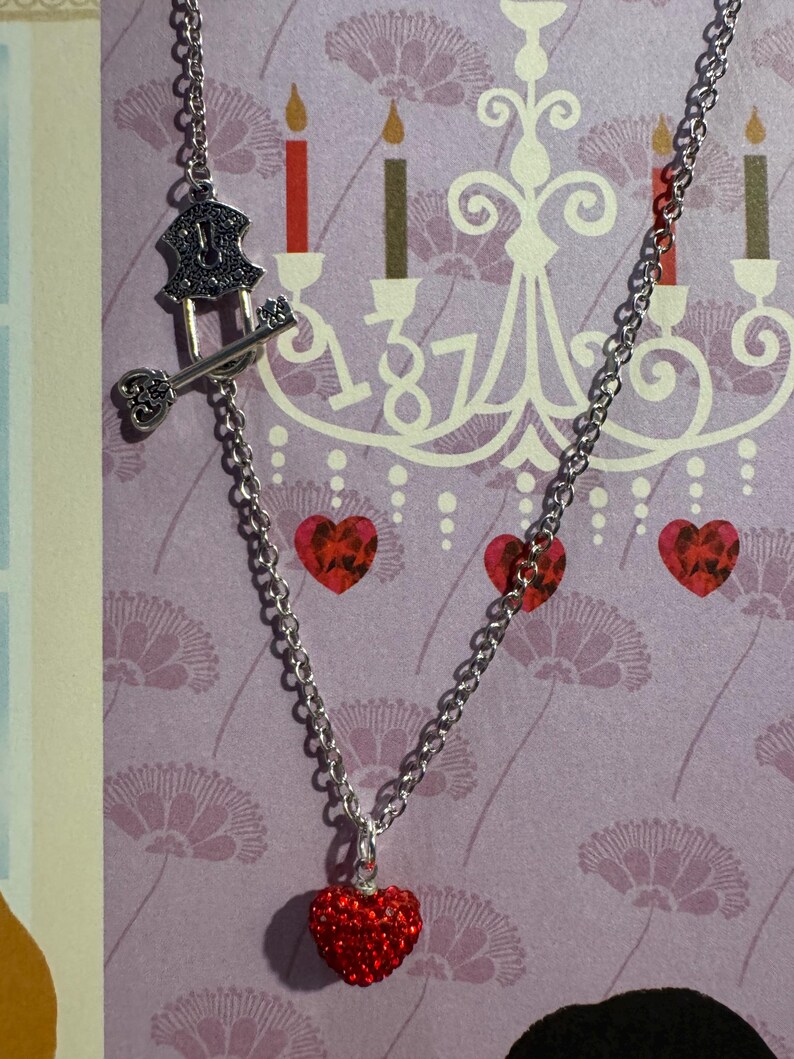 May include: A silver chain necklace with a red heart pendant. The necklace also features a key and lock charm. The background is a purple floral pattern with a chandelier and candles.