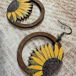 May include: Wooden hoop earrings featuring a sunflower design. The earrings have a dark brown wooden frame with a yellow and brown sunflower on the lower half. The earrings have a hook closure.