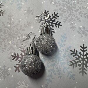 May include: A pair of silver glitter ball earrings. The earrings are designed to resemble miniature Christmas ornaments, with a silver hook and a sparkly, textured surface. The earrings are set against a background with snowflake patterns.