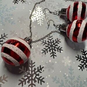 May include: Red and white striped Christmas ornament earrings. The earrings are made of small, round ornaments with silver-colored metal findings and chains. The background is a light gray paper with snowflake patterns.