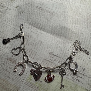 Puede incluir: Una pulsera de plata con varios dijes, incluyendo una guitarra, un coraz&oacute;n, una herradura, un meg&aacute;fono con el texto "CHEERS", un bal&oacute;n de f&uacute;tbol americano, una llave y un anillo. La pulsera se muestra sobre un fondo de peri&oacute;dico vintage.
