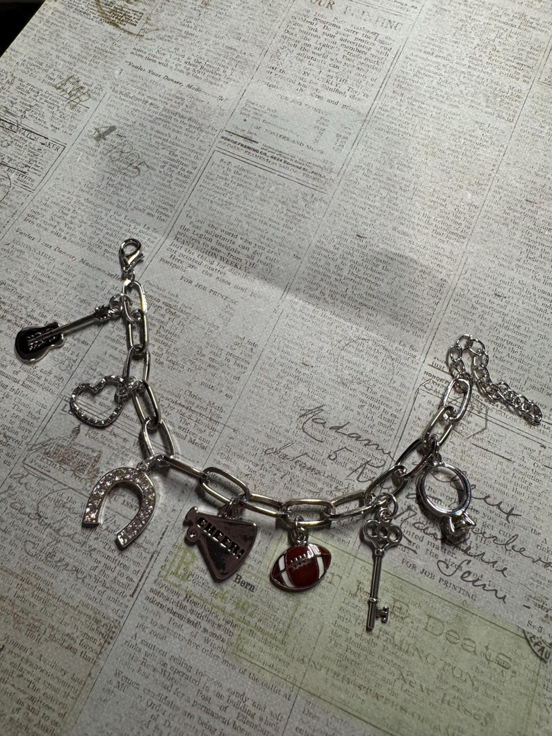 Puede incluir: Una pulsera de plata con varios dijes, incluyendo una guitarra, un coraz&oacute;n, una herradura, un meg&aacute;fono con el texto "CHEERS", un bal&oacute;n de f&uacute;tbol americano, una llave y un anillo. La pulsera se muestra sobre un fondo de peri&oacute;dico vintage.