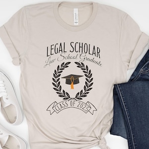 May include: A beige t-shirt with the text "Legal Scholar Law School Graduate Class of 2025" and a black graduation cap with a gold tassel inside a laurel wreath.