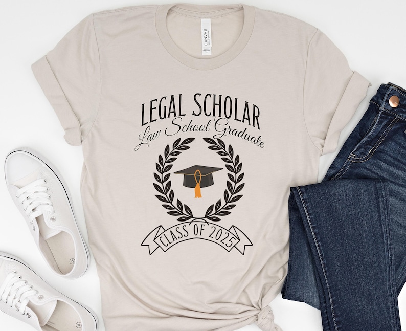 May include: A beige t-shirt with the text "Legal Scholar Law School Graduate Class of 2025" and a black graduation cap with a gold tassel inside a laurel wreath.