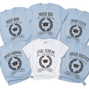 May include: Six light blue t-shirts with black text and graphics. The shirts say "Proud Mom", "Proud Grandma", "Proud Dad", "Proud Sister", "Proud Brother", and "Legal Scholar". Each shirt has a laurel wreath with a graduation cap and the text "Law School Graduate" and "Class of 2023".