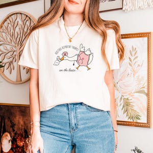 May include: A white t-shirt with a graphic of a goose wearing a nurse's uniform and holding a tissue box. The text on the shirt reads "Registered Respiratory Therapist on the loose..."