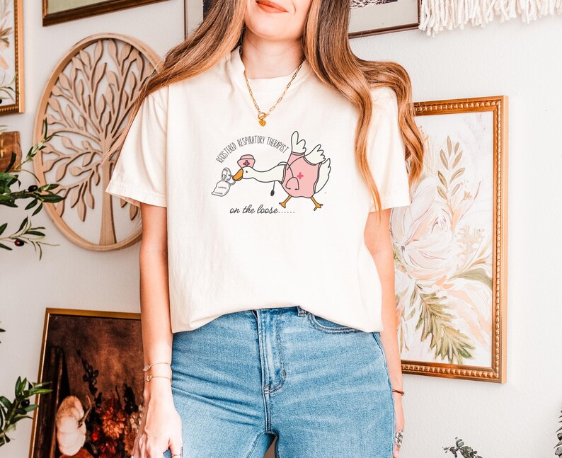 May include: A white t-shirt with a graphic of a goose wearing a nurse's uniform and holding a tissue box. The text on the shirt reads "Registered Respiratory Therapist on the loose..."