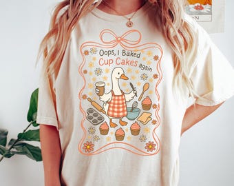 May include: Cream-colored t-shirt featuring a cartoon duck wearing a red and white checkered apron, holding a coffee cup and a spoon. The text "Oops, I Baked Cup Cakes again" is displayed within a floral frame.
