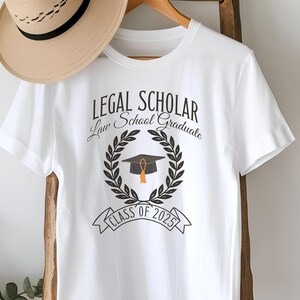 May include: White t-shirt with the text "LEGAL SCHOLAR Law School Graduate Class of 2025" and a black graduation cap with a gold tassel inside a laurel wreath.