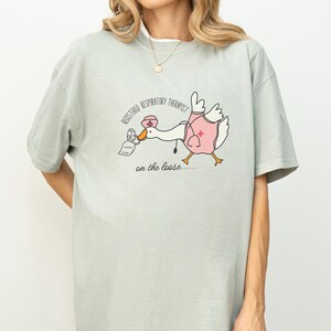 May include: A light green t-shirt with a cartoon goose wearing a nurse's hat and holding a nebulizer. The text on the shirt reads "Registered Respiratory Therapist on the loose..."