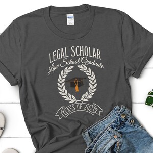 May include: A dark gray t-shirt with the text "LEGAL SCHOLAR Law School Graduate Class of 2025" and a graduation cap with a tassel in a wreath.