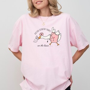 May include: Pink t-shirt with a cartoon goose wearing a nurse's hat and holding a face mask. The text on the shirt reads "Registered Respiratory Therapist on the loose..."