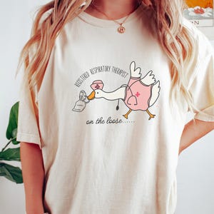 May include: A white t-shirt with a cartoon goose wearing a nurse's hat and holding a face mask. The text on the shirt reads "Registered Respiratory Therapist on the loose..."