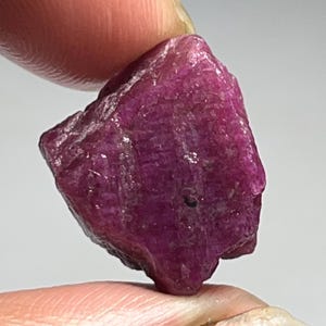 May include: A rough, uncut ruby gemstone with a deep red-purple hue. The raw mineral is held between two fingers, showcasing its natural, irregular shape and textured surface. The gemstone is a vibrant example of a natural mineral.