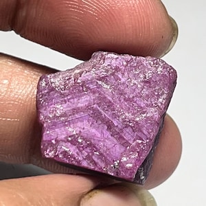 May include: A rough-cut, natural ruby gemstone. The ruby is held between fingers and has a deep pinkish-red color with visible crystalline structures. The surface has a textured appearance, with light reflecting off the facets.