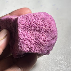 May include: A close-up of a porous, cube-shaped object in a vibrant shade of purple. The object has a textured surface with numerous small holes, and is held in a person's hand.