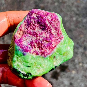Pode incluir: Uma gema em bruto com um contraste de cores vibrante. O centro apresenta um tom magenta profundo, rodeado por um exterior verde brilhante. A textura da pedra é áspera, mostrando sua forma natural.
