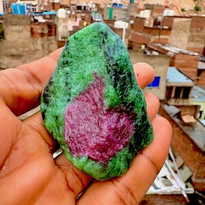 May include: A raw, unpolished gemstone featuring a vibrant magenta-colored center surrounded by a textured, mottled green and black mineral matrix. The stone has an irregular, angular shape with visible crystalline structures and natural surface variations.