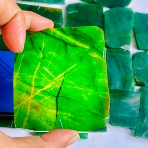 Puede incluir: Una mano sostiene una vibrante piedra de jade verde con vetas amarillas y una grieta prominente. La piedra es rectangular y pulida, con una superficie brillante. Otras piezas de jade similares están en el fondo.