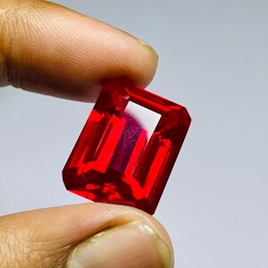May include: A vibrant, transparent red gemstone with a rectangular emerald cut, featuring sharp, clean facets that reflect light to create a brilliant, deep crimson appearance throughout the stone's structure and polished surface.