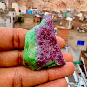 May include: A raw, unpolished gemstone specimen featuring a distinct colour combination of vibrant magenta-purple and bright lime-green. The stone has an irregular, jagged shape with a rough, textured surface, showcasing natural mineral formations.