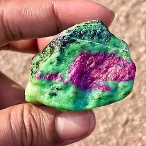 May include: A natural gemstone with a striking colour palette. The stone showcases a blend of bright green, deep magenta, and black. The magenta is centrally located, the green forms the base, and black accents the edges. A unique mineral specimen.