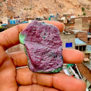 May include: A raw, unpolished mineral specimen featuring a prominent, textured deep magenta and purple center with distinct patches of opaque, light green material along the edges. The stone has an irregular, jagged shape with a rough surface.
