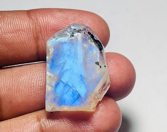 May include: A raw, uncut moonstone gemstone with a vibrant blue flash. The stone has a rough, natural texture and a translucent quality, with areas of white and black. The gemstone is held in a hand.