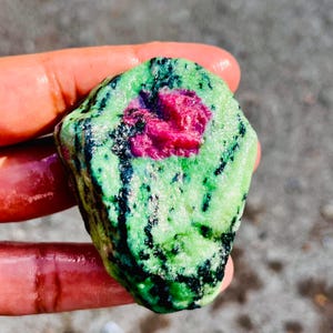 Könnte beinhalten: Ein naturbelassener Edelstein mit einem leuchtend roten Kern, der von einer gesprenkelten grün-schwarzen Oberfläche umgeben ist. Die unregelmäßige Form des Steins unterstreicht seine rohe Schönheit.