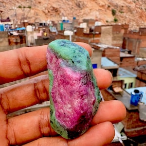 May include: A raw, unpolished gemstone featuring a vibrant magenta-pink center surrounded by mottled green and black mineral deposits. The stone has an irregular, elongated shape with a textured, crystalline surface and natural color variations.