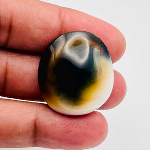 May include: A smooth, oval-shaped cabochon gemstone featuring a gradient of colors. The center is a deep, opaque black that transitions into a warm, golden-yellow hue, eventually fading into a creamy white border along the outer edge of the stone.